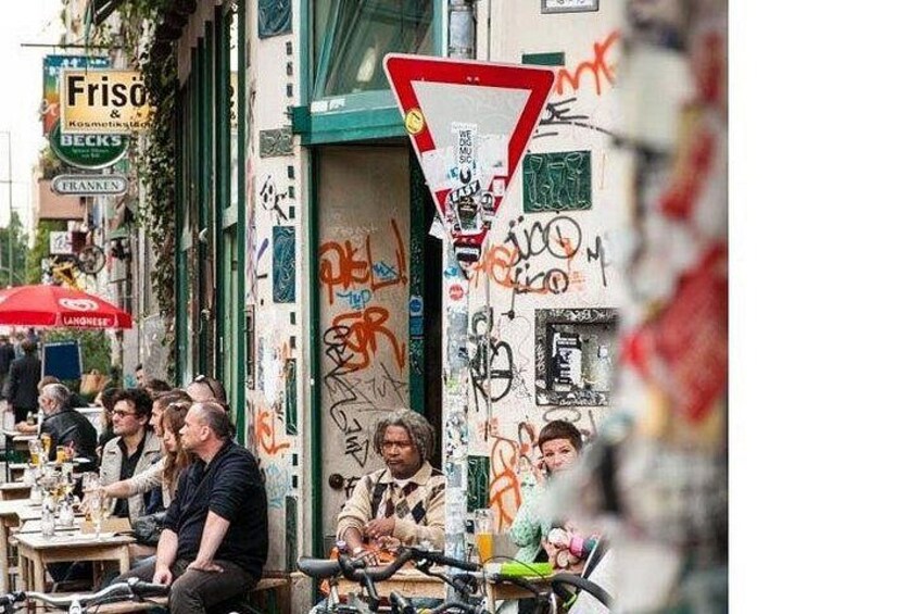 Straßenszene mit Personen, die an Tischen vor einem Café sitzen, einem roten Stoppschild und Graffiti an den Wänden. 5