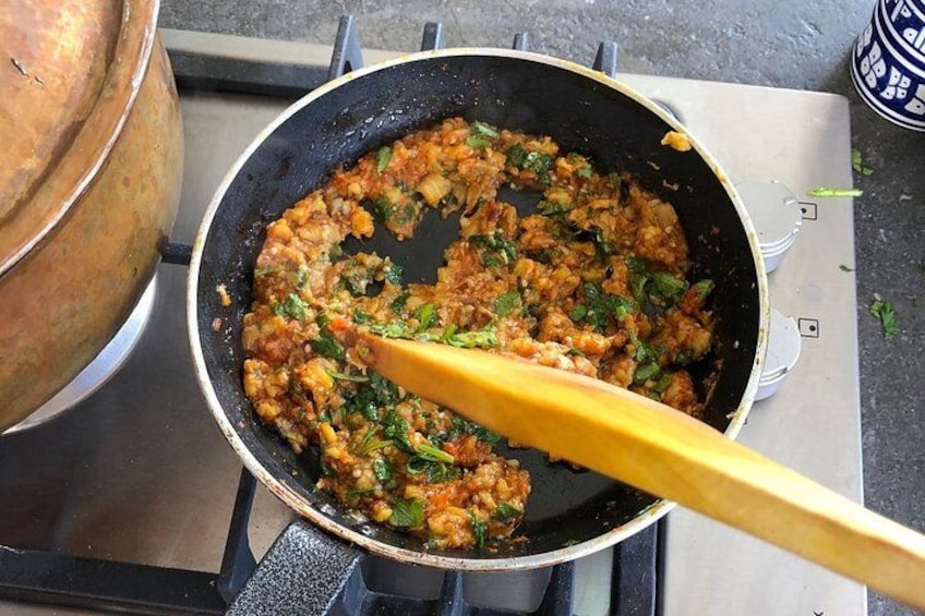 Eine Bratpfanne auf einem Herd, Essen wird zubereitet, ein Holzlöffel und im Hintergrund ein großer Kupfertopf. 2