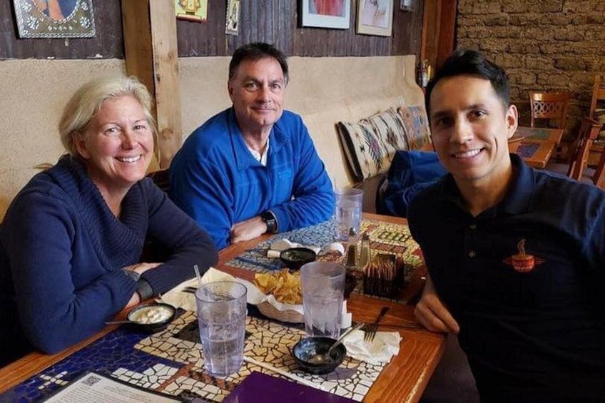 Three people seated at a table with a mosaic tablecloth, a plate of food, and a glass of water. 4