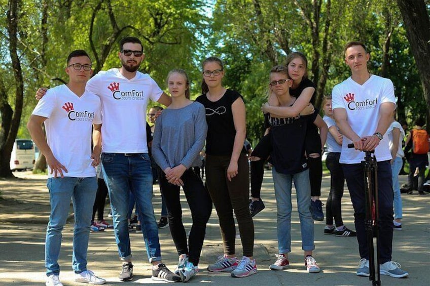 A group of people standing outdoors, some wearing white t-shirts with a logo, in a park-like setting with trees and a paved path. 2