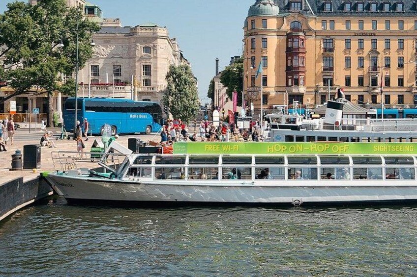 A riverboat with 'FREE WI-FI' and 'SIGHTSEEING' signs, docked near a city with buildings and a blue bus in the background. 3