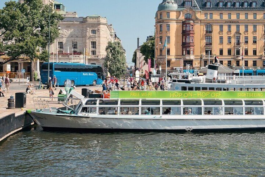 Un bateau de tourisme navigue sur une rivière, transportant des passagers. On aperçoit également un autobus bleu et des bâtiments en arrière-plan. 5