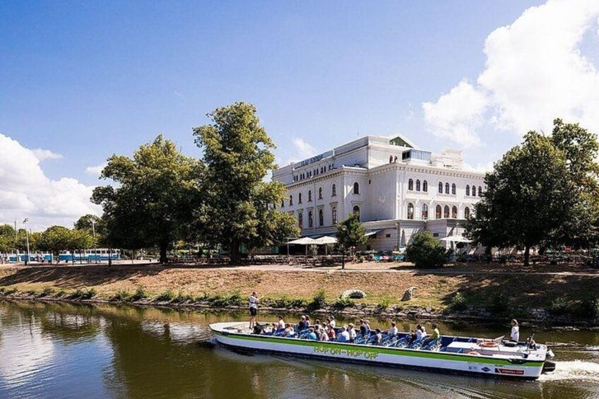 A river with a boat carrying passengers, a large white building with arched windows, and trees along the riverbank. 5