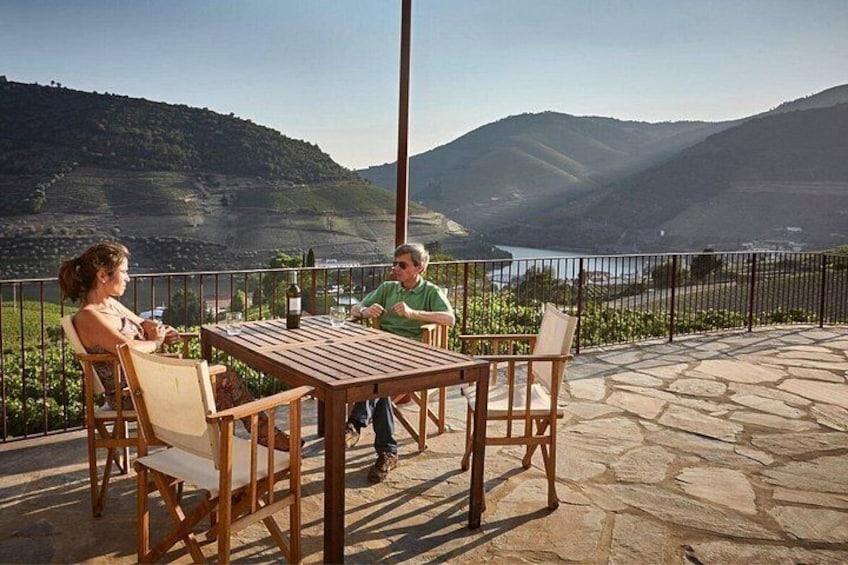 A wooden table and chairs on a stone patio overlooking a valley with a river and hills. 3