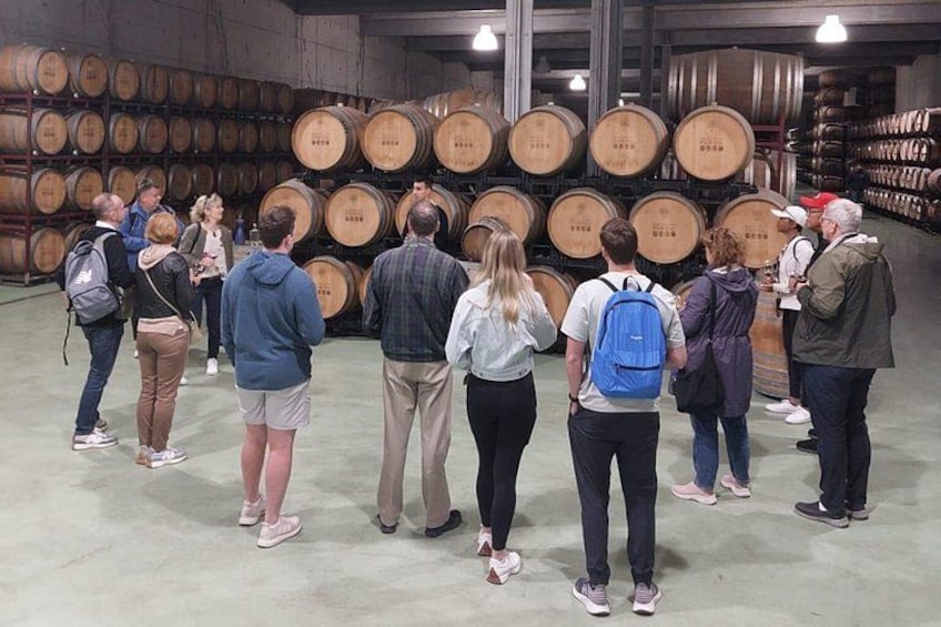 A group of people standing in a warehouse with rows of wooden barrels. 4