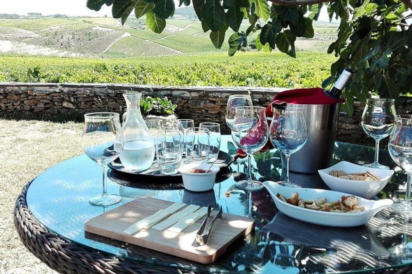 Une table à manger installée à l’extérieur, avec des verres à vin, une carafe et une bouteille de vin, le tout mis en valeur par un paysage de vignes en arrière-plan. 1