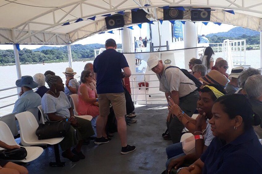 Een groep mensen op een boot, sommigen staan en sommigen zitten, met een bergachtig landschap op de achtergrond. 3