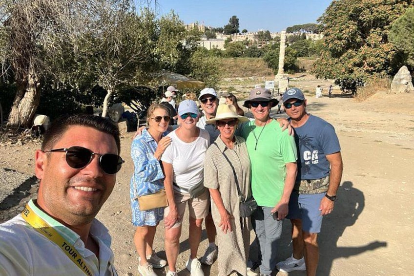 A group of people standing outdoors, some wearing hats and sunglasses, with trees and a clear sky in the background. 5