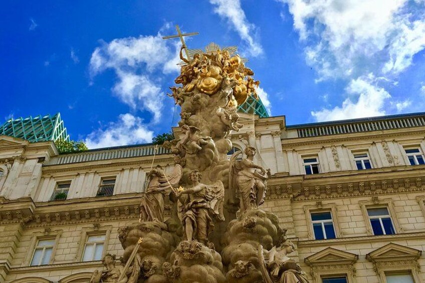 A classical statue on a pedestal with a cross, set against a building with windows and a blue sky with clouds. 5