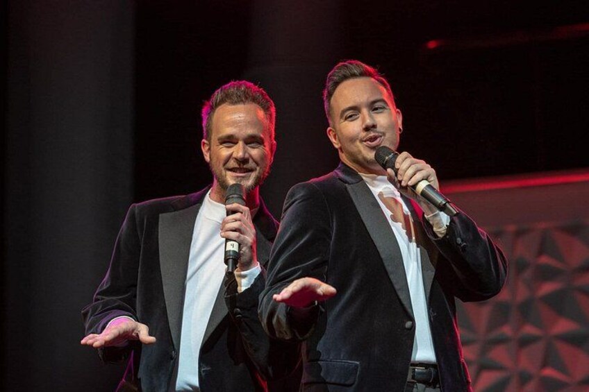 Deux hommes vêtus de costumes noirs, tenant des microphones sur une scène dont le fond est orné d’un motif géométrique. 4