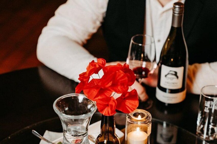 Un tavolo con una bottiglia di vino, un bicchiere, una candela e un vaso con fiori rossi. 2