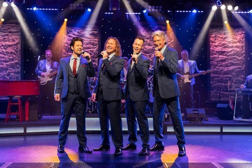 A stage with four men singing, a keyboard, and a band in the background. 1