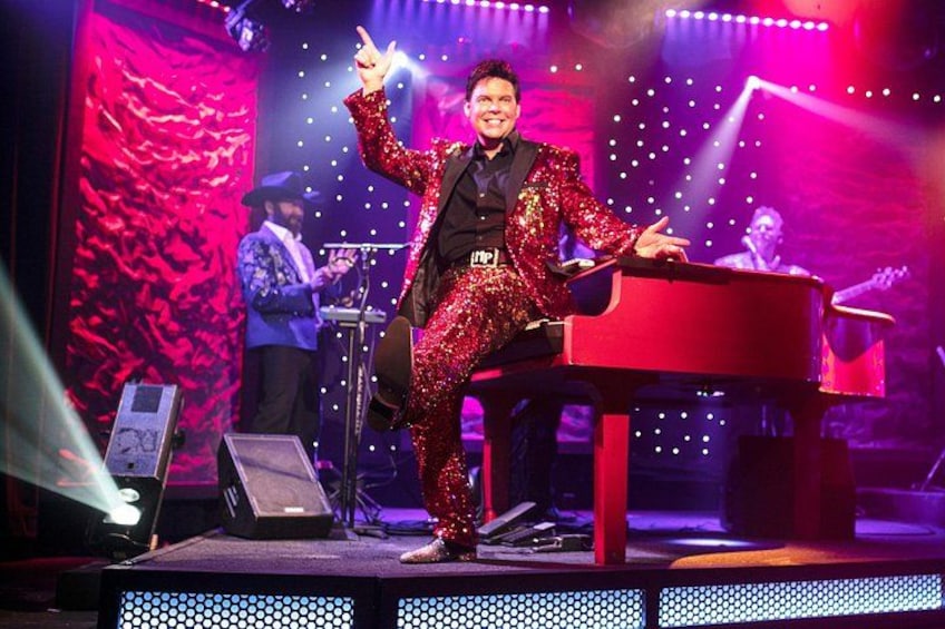 A person in a sequined outfit performing on stage with a red piano, a large screen in the background, and stage lighting. 4