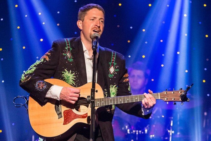 En person spiller akustisk guitar på en scene med en mikrofon, iført en sort jakke med farverigt broderi. 4