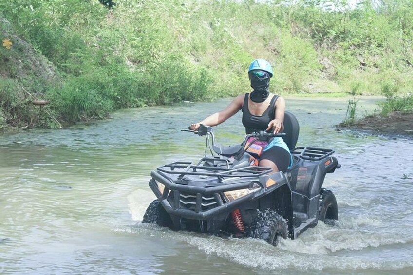 A person on an ATV navigating through a river surrounded by greenery. 1