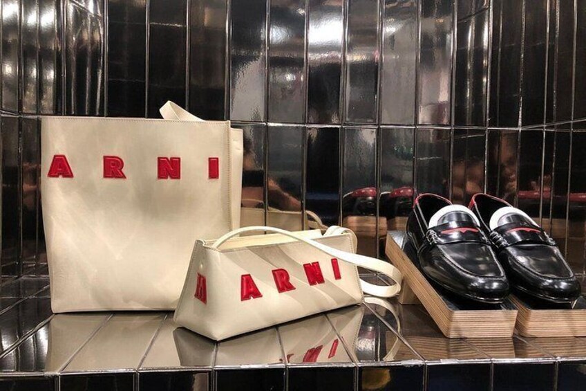 Eine mit Fliesen verkleidete Wand mit glänzender Oberfläche, eine weiße Handtasche mit dem Schriftzug 'ARNI' und schwarze Schuhe. 3