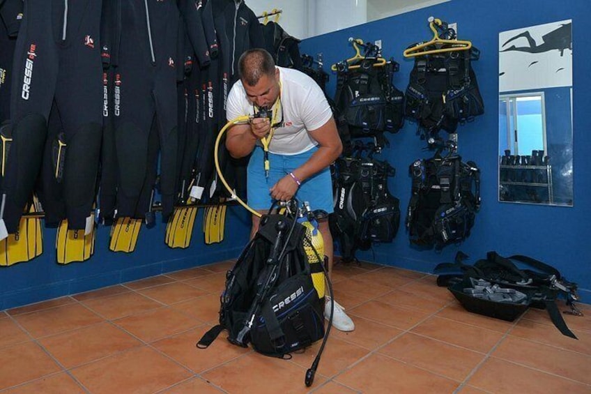 A person adjusting a scuba diving backpack in a room with multiple diving suits and equipment. 1