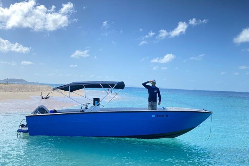 A blue boat on calm turquoise waters near a sandy beach with a person standing on the deck. 4