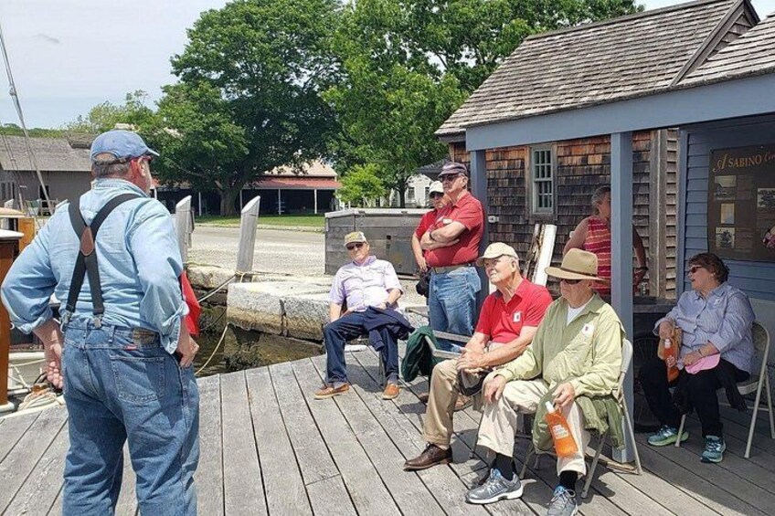 A group of people on a wooden dock, some standing and some sitting, with a small building in the background. 3