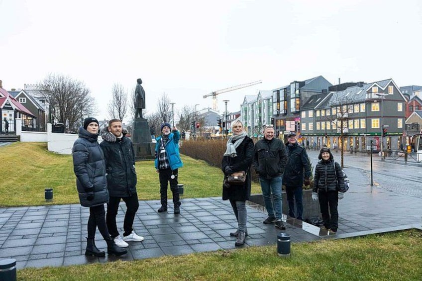 A group of people standing on a paved area with a statue in the background, surrounded by buildings and a grassy area. 2