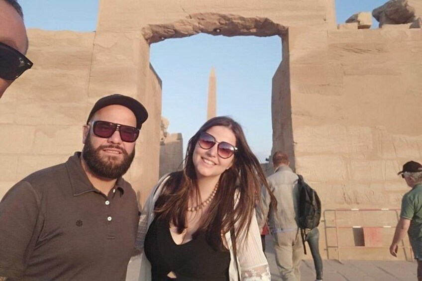 Un homme et une femme se tiennent devant une structure de l’Égypte ancienne, avec une grande arche en pierre et un obélisque imposant en arrière-plan. 3