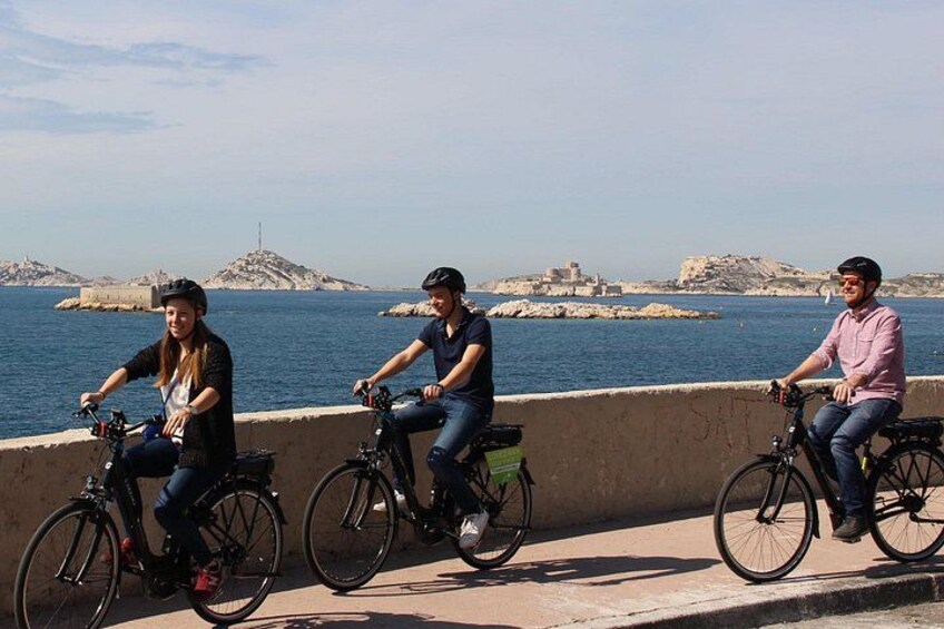 Trois personnes font du vélo sur une piste qui longe le bord de l’eau, avec des affleurements rocheux et une île lointaine en arrière-plan. 1