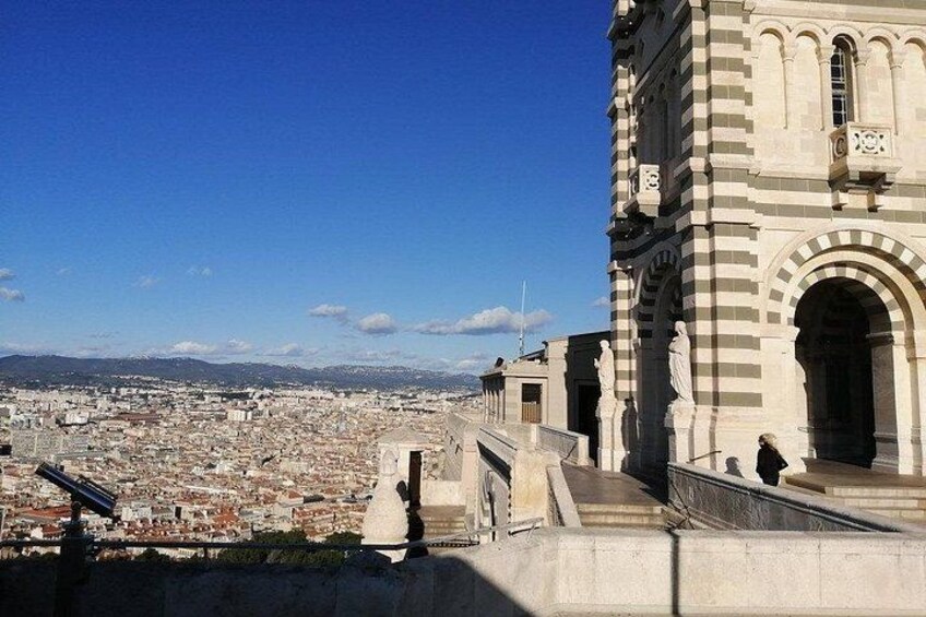 Une vue urbaine mettant en valeur un bâtiment imposant, orné d’arches et de statues, qui surplombe une zone densément peuplée sous un ciel dégagé. 3