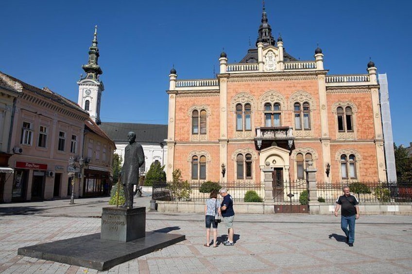 A historical building with a statue in front, a church with a spire in the background, and people walking. 5