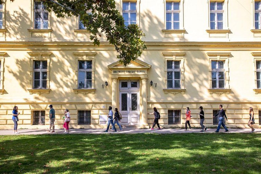 A building with a classical facade, a tree in front, and people walking on the sidewalk. 3