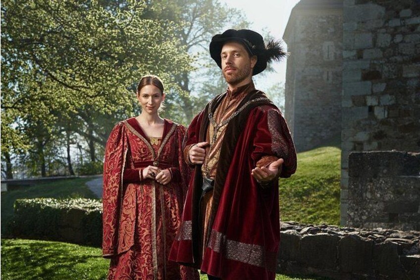 Two people in historical costumes standing in a park with a stone structure in the background. 1
