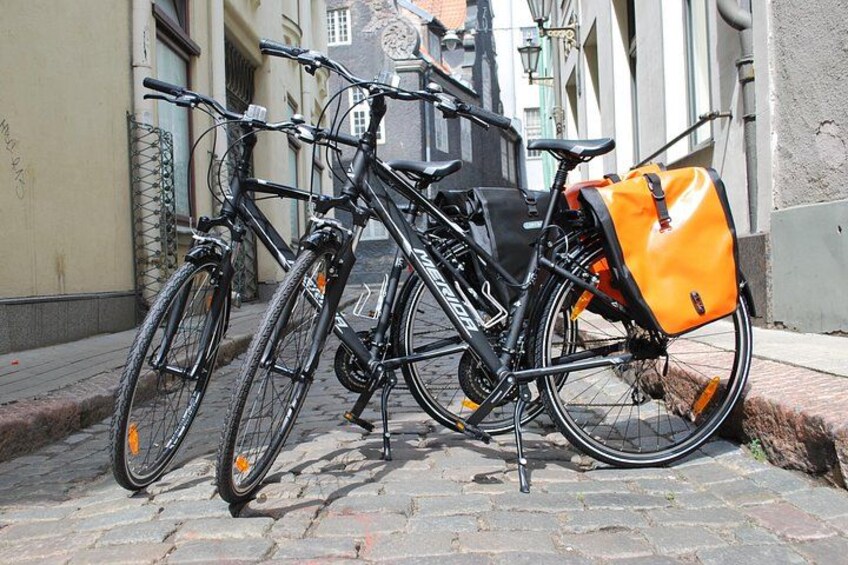 Twee fietsen geparkeerd op een geplaveid straatje, waarvan er een een oranje tas aan de achterkant heeft. 1
