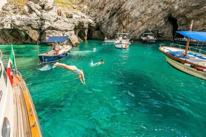 A clear turquoise sea with boats, rocky cliffs, and a person diving. 1