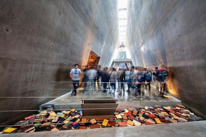 A group of people standing in a large, open, concrete structure with a pile of books in the foreground. 2