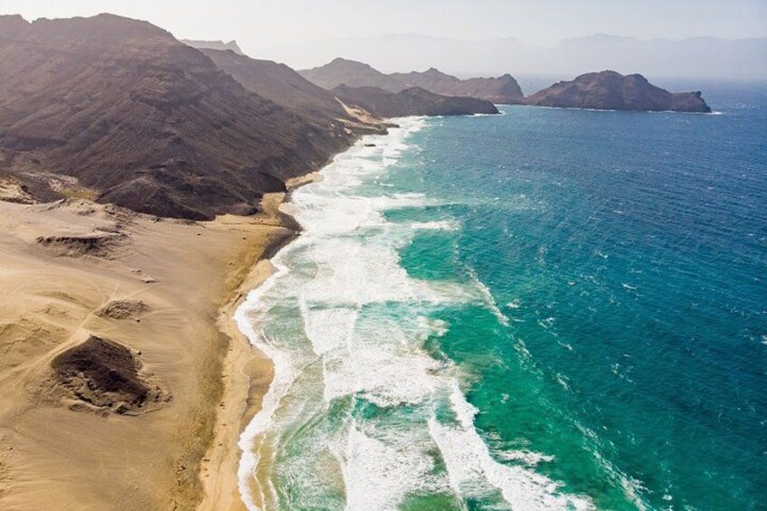 A coastal landscape with a sandy beach, turquoise ocean waves, and distant mountains. 3