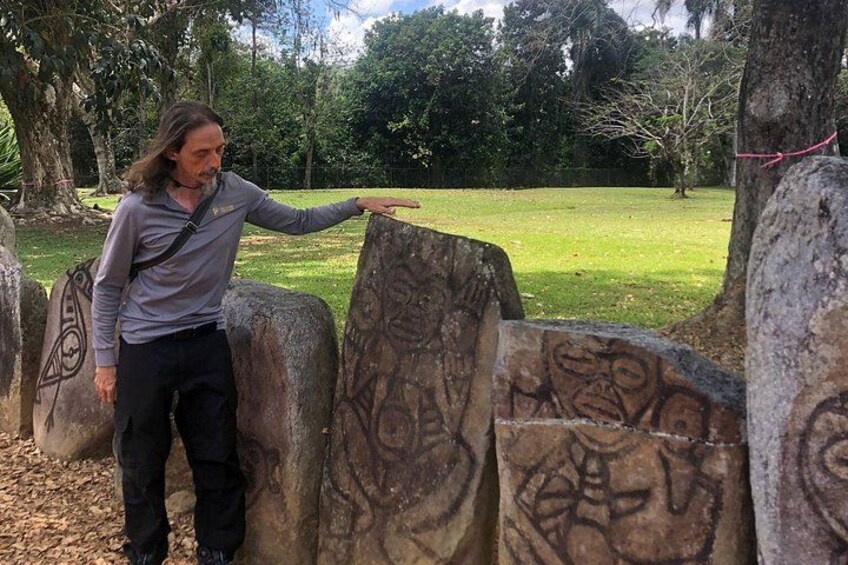 A person standing next to large carved stones with intricate designs, in a grassy area with trees in the background. 2