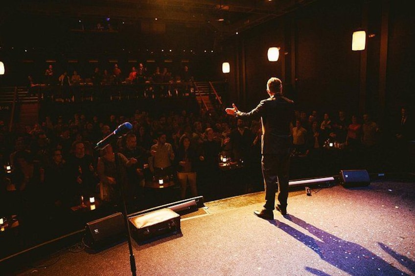 Una persona sul palco con un microfono, che si rivolge a un pubblico in un auditorium poco illuminato. 4
