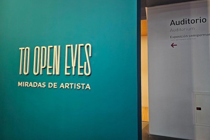 Una parete color petrolio con il testo 'TO OPEN EYES MIRADAS DE ARTISTA' e una segnaletica per 'Auditorio Exposición semipermanente'. 2