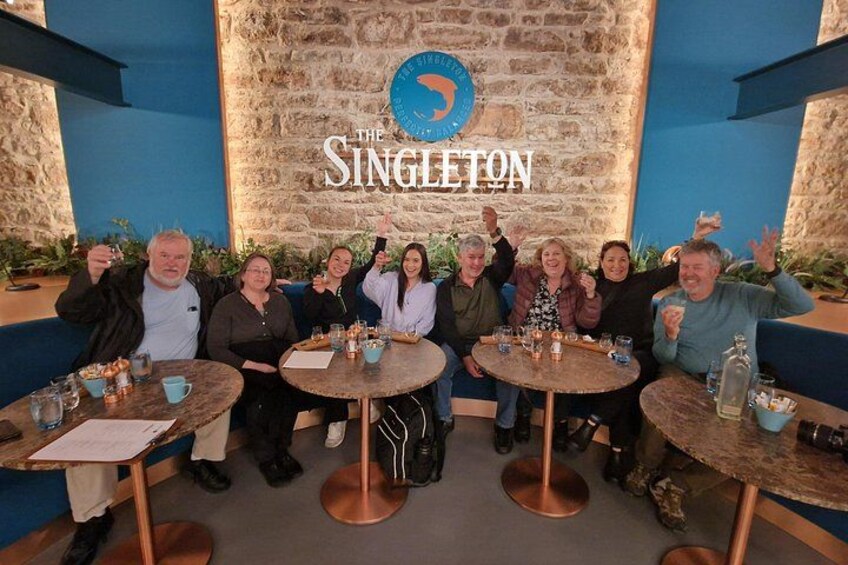 A group of people seated at round tables with drinks, in a restaurant with a stone wall and a sign reading 'The Singleton'. 4