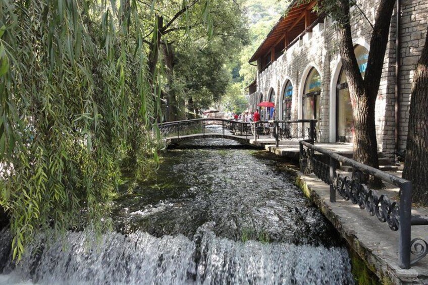 A stone bridge over a flowing stream, surrounded by trees and a building with arched windows. 2