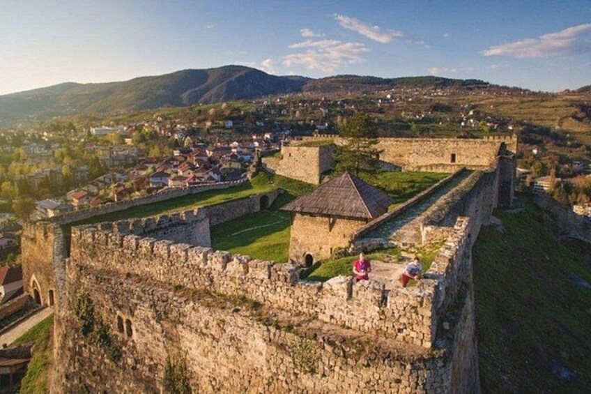 A historic stone fortress with a moat, surrounded by a town and hills in the background. 1
