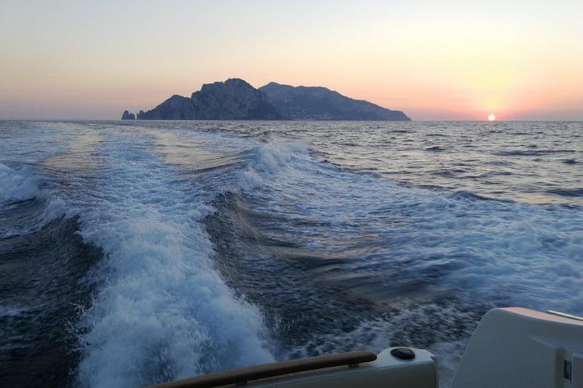 Ein Boot bewegt sich durch unruhiges Wasser, im Vordergrund sind Wellen zu sehen. Im Hintergrund eine ferne Insel mit einem Berggipfel und der untergehenden Sonne am Horizont. 4