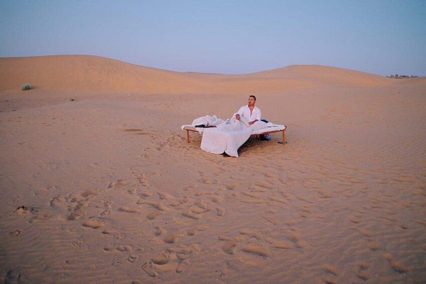 Eine Person sitzt auf einem Bett, im Hintergrund Dünen in einer Wüstenlandschaft. 2