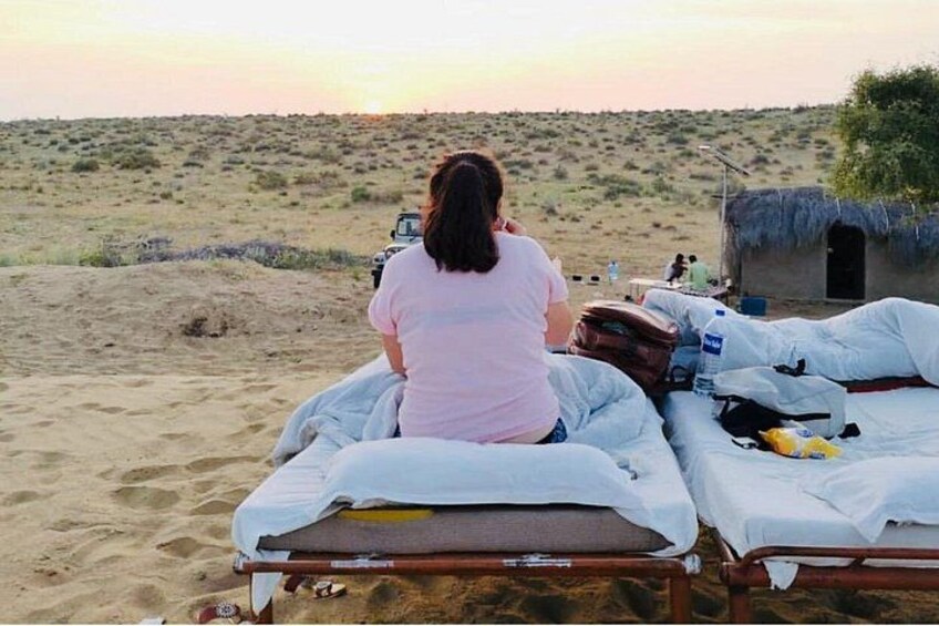 Eine Person sitzt auf einem Bett in einer Wüstenlandschaft mit Sanddünen und einer kleinen Hütte im Hintergrund. 1