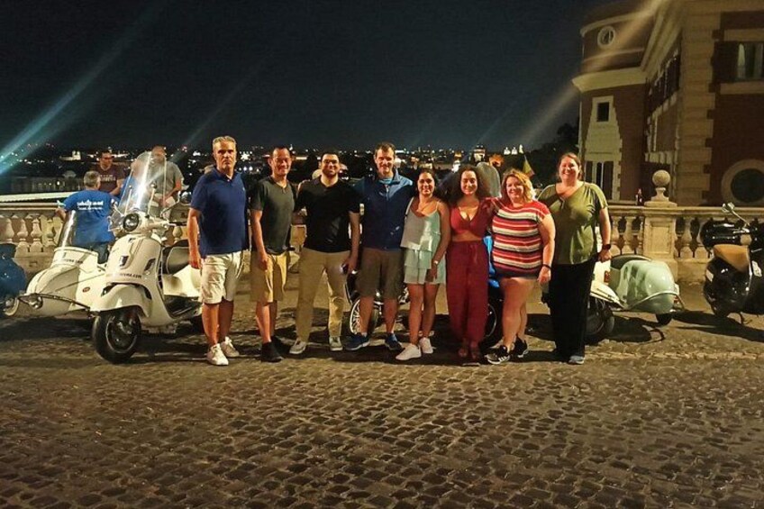 A group of people standing on a cobblestone street at night with a scooter and a motorcycle in the background. 2