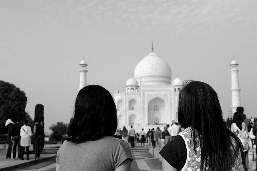 Two people facing the Taj Mahal, a large white marble mausoleum with domes and minarets. 2