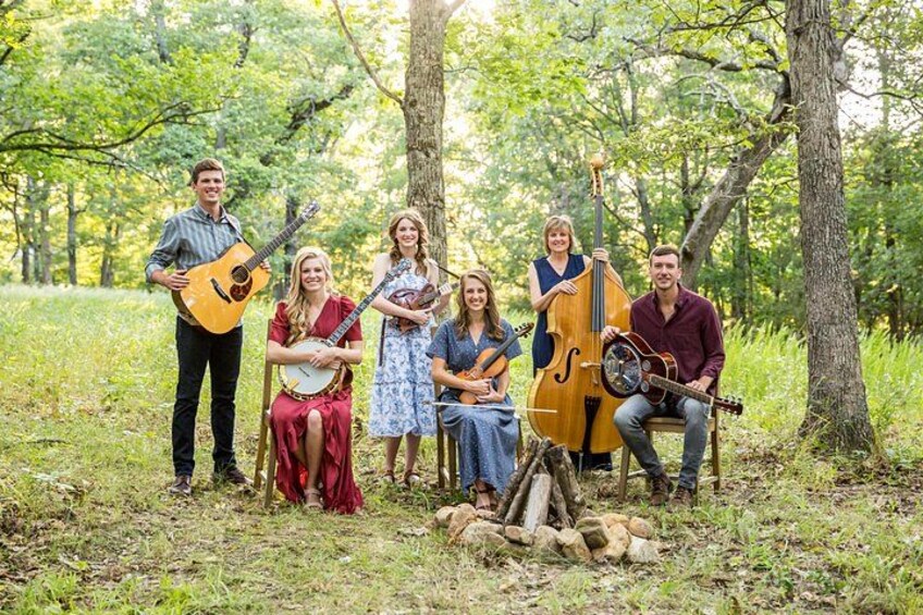 A group of people in a wooded area with trees and grass, some holding musical instruments. 3