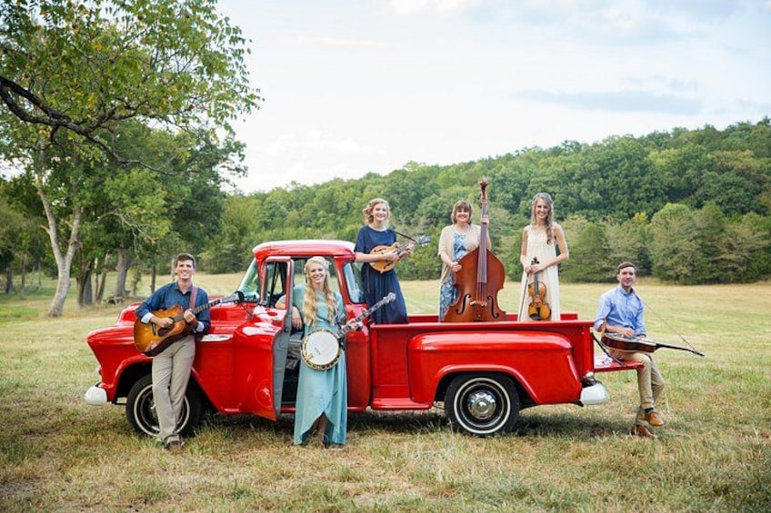Een groep mensen met muziekinstrumenten in een rode pick-up truck op een grasveld, met bomen op de achtergrond. 2