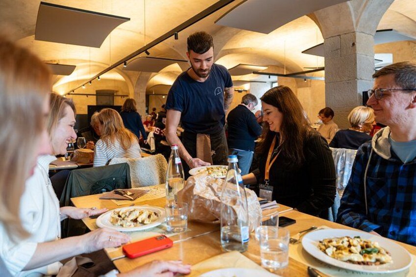 A restaurant with people dining, tables with plates and glasses, and a waiter serving food. 4