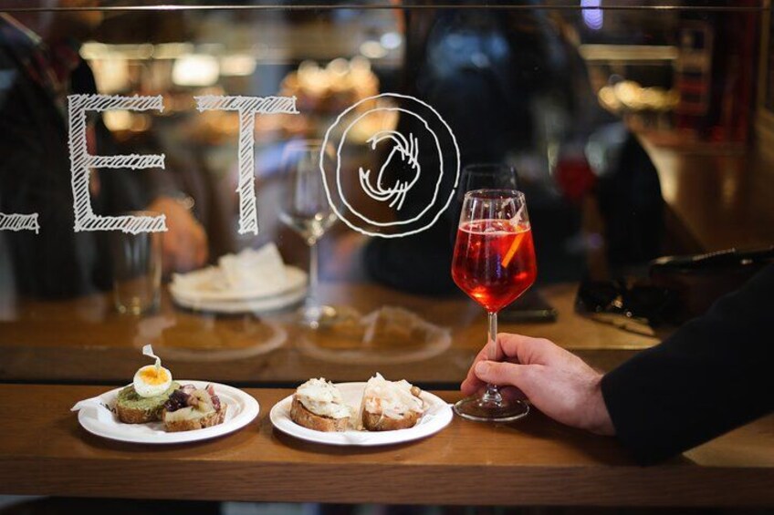 Una persona sosteniendo una copa de vino tinto en un bar, con platos de comida al fondo. 1