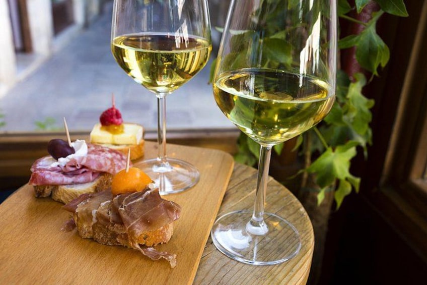 Une table en bois avec un verre de vin blanc, une planche en bois sur laquelle sont disposés divers fromages et différentes sortes de charcuterie, et une fenêtre donnant sur un paysage verdoyant. 2